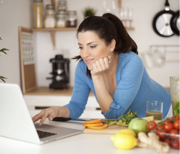 Woman in kitchen using a laptop, surrounded by fresh fruits and vegetables on the counter.