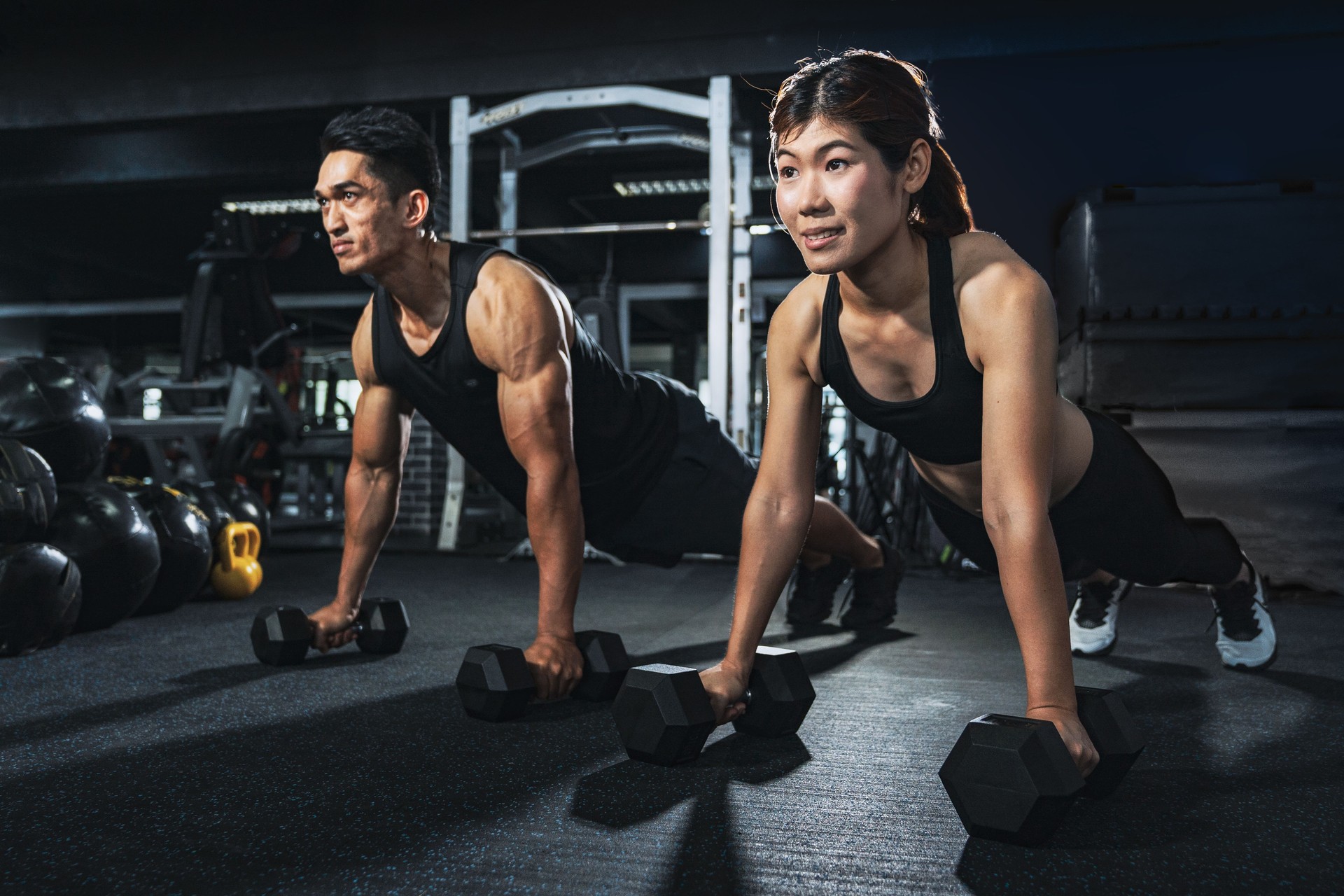 Young couple is working out at gym. Attractive woman and handsome muscular man are training in light modern gym. Doing plank on kettlebell. Push-up on weights.